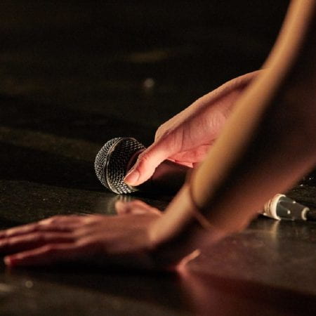 MA Theatre Artist Bekki Loveridge hands picking a microphone up off the stage floor as part of her performance work.