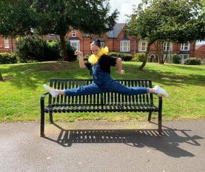 A performer in a yellow net collar and dungarees performs a splits on a park bench.