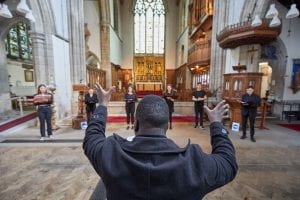 A conductor for UoL BA Music stand with his back to the camera and directs musicians stood in the nave of an elaborately decorated church.