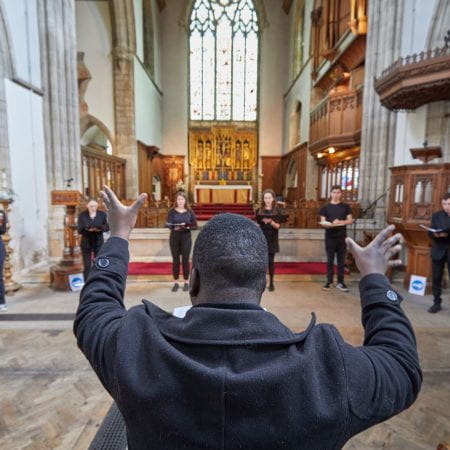 A conductor for UoL BA Music stand with his back to the camera and gestures to musicians stood in the nave of an elaborately decorated church.