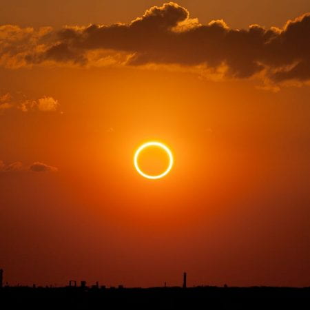 A rare 'ring of fire' solar eclipse. The sun appears as a golden ring in a flame-oramge sky. Clouds gather overhead.
