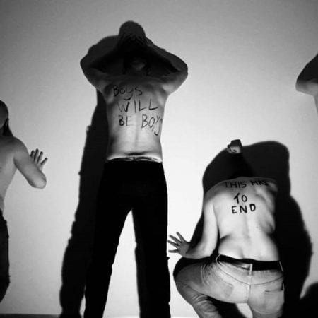 The Unbeatable Foe Promo Image: A black and white photo - 4 men in dark trousers leaned up against a wall with hands against it. From left to right, the first man is leaning forward into the wall, the second is stood with hands against the wall, the third is crouched and the fourth is also stood. The writing on their naked backs in black ink reads “Grow some balls”, “Boys will be Boys”, “This has to End”, and “Man Up”.