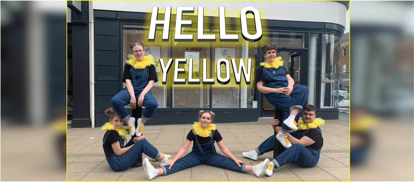 Five performers (four female and one male) posed in front of a large sign which reads 'HELLO YELLOW'. The performers are in various positions in the foreground and background, sitting and standing on bollards outside a building. All performers are looking at the camera, wearing blue dungarees, a black t-shirt and yellow netted ruffle around their neck.