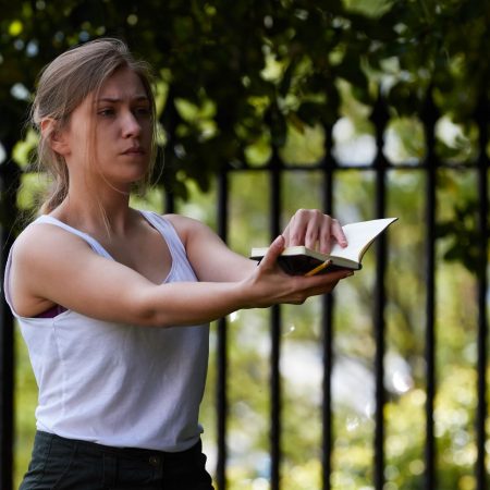A performer in a white vest and with long hair tied back in a ponytail, is stood outside infron of trees and railings, holding a small notebook in their outstretched hands.
