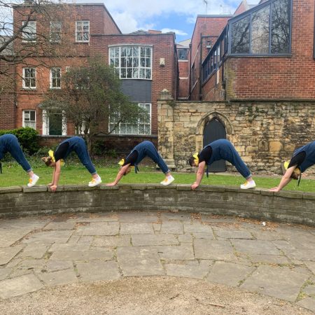 Five performers in dungarees perform an 'on all fours' elephant walk along a low stone wall.