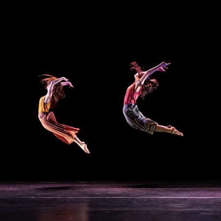 Two dancers in red and orange clothing jump high in the air, their arms extended above their heads and backs bending in an arch.