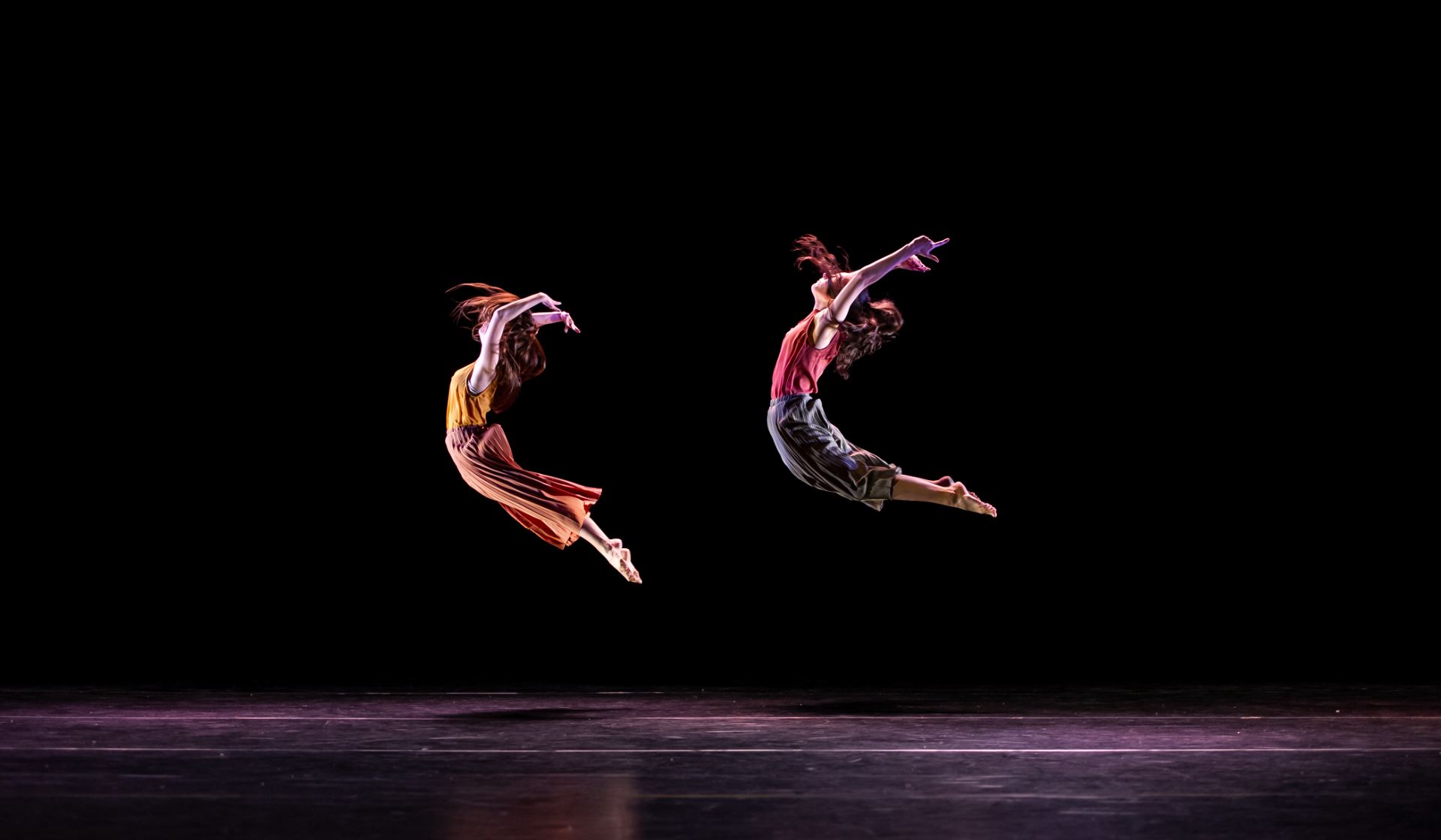 Two dancers in red and orange clothing jump high in the air, their arms extended above their heads and backs bending in an arch. 